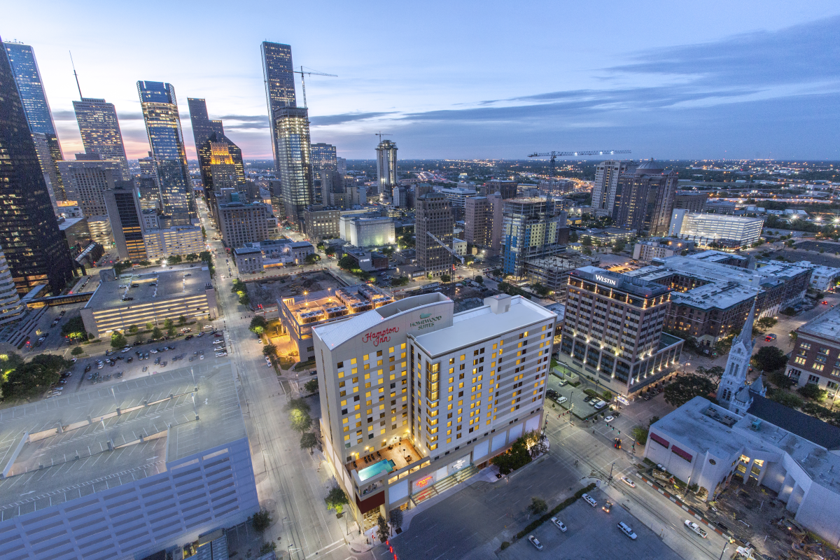 Aerial view of a bustling cityscape at dusk, featuring tall, illuminated skyscrapers and a brightly lit Hampton Inn in the foreground.