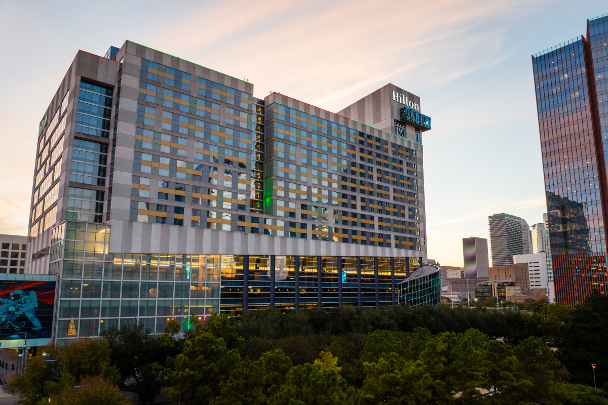 A modern hotel building with reflective glass windows and a "Hilton" sign is set against a cityscape at sunset, surrounded by trees.