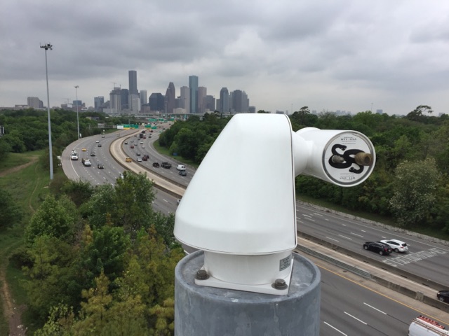A white traffic camera is mounted on a pole, overlooking a highway with moving cars. The Houston skyline is visible in the background under a cloudy sky.