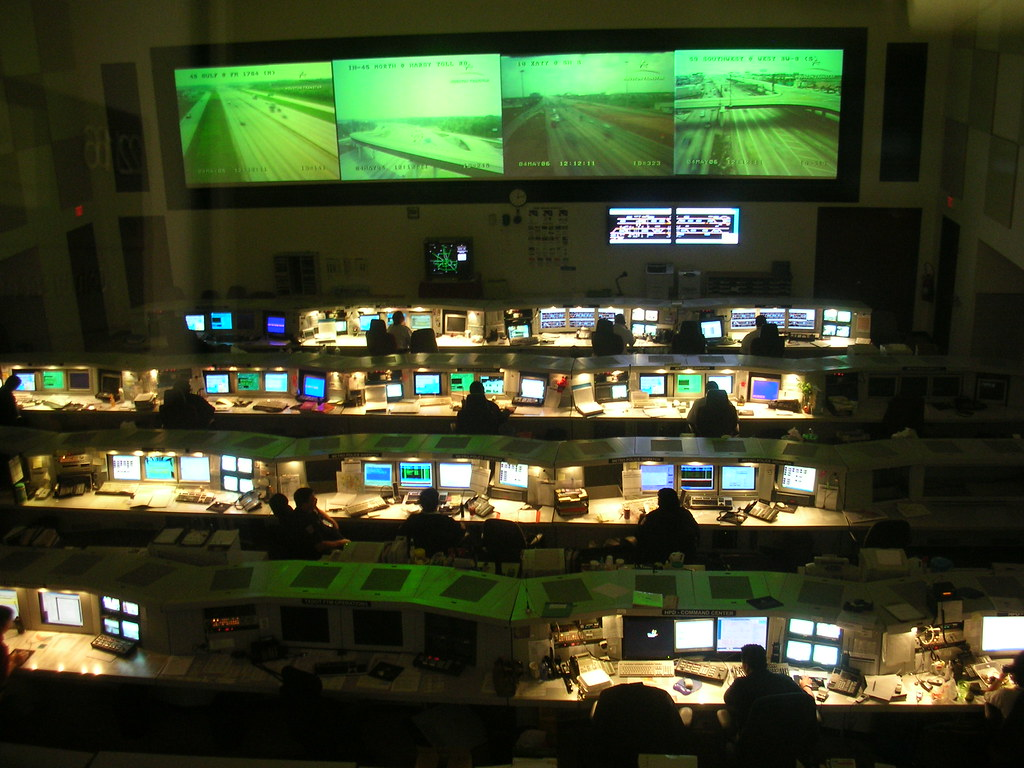 Dimly lit control room with multiple workstations and computer screens. Large overhead monitors display green-tinted images.