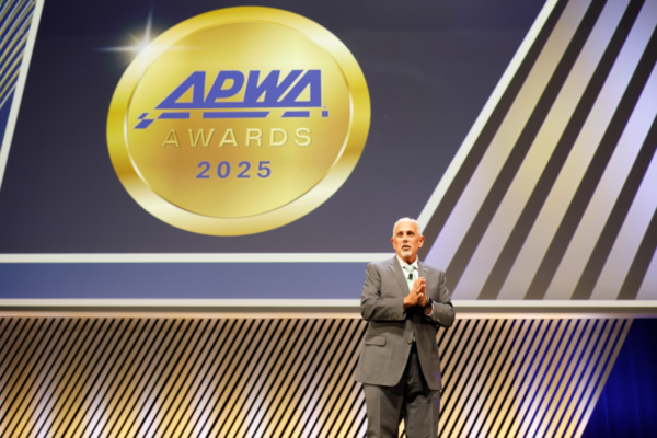 A man in a suit stands on stage at the APWA Awards and Recognition Ceremony at PWX. Behind him is a large screen with a gold logo and diagonal stripes.