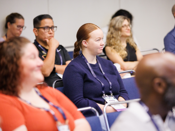 PWX attendees sit attentively during an education session. A woman in a navy sweater is focused, surrounded by others listening intently.