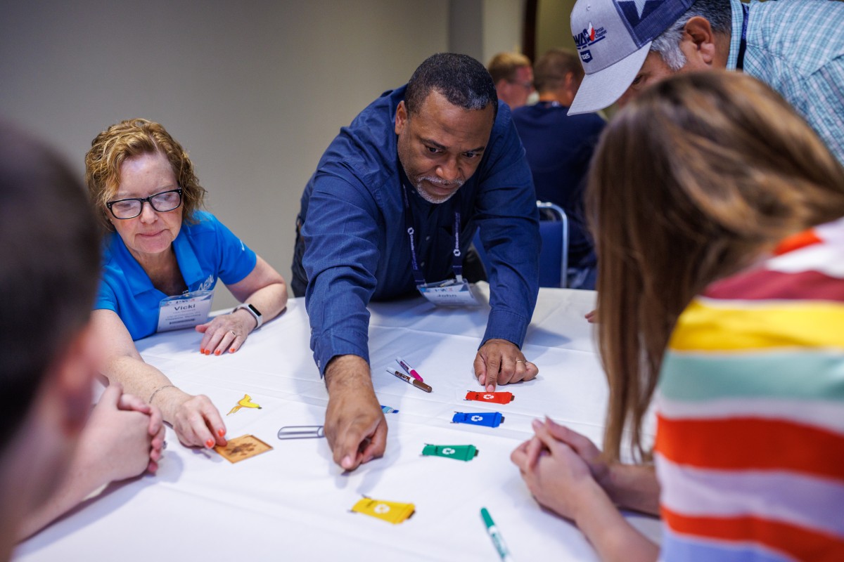 A diverse group of people gathers around a table engaged in a collaborative game as part of an escape room at PWX.