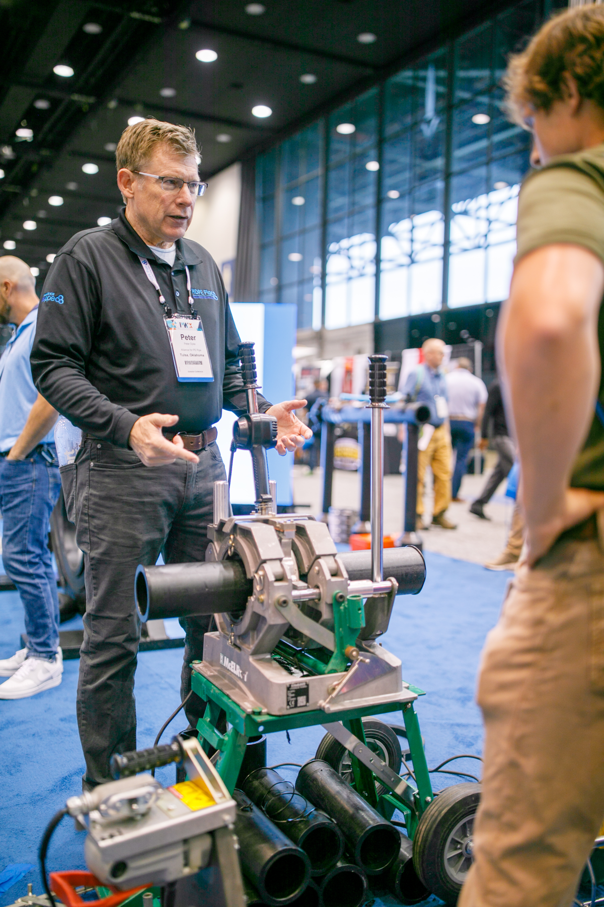 A man in a black shirt and name badge explains a machine in the PWX exhibit hall.