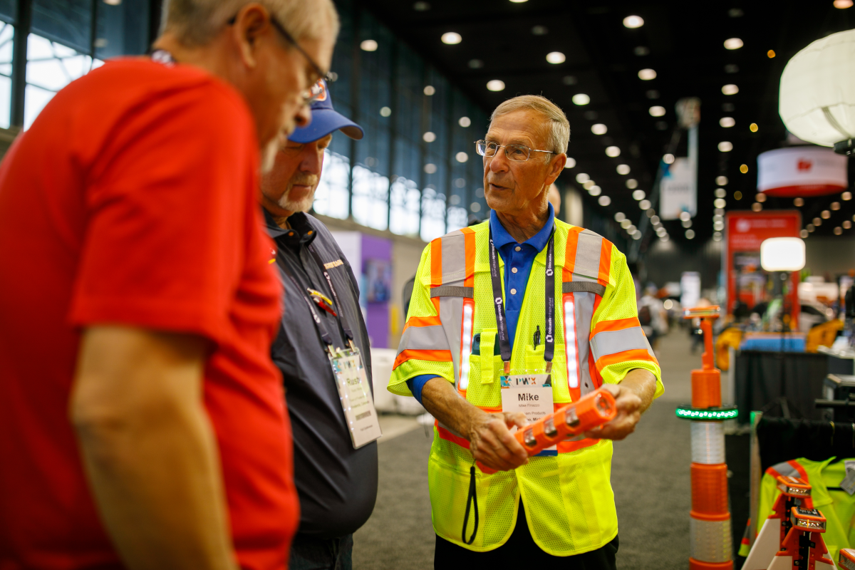 A man in a neon safety vest demonstrates a tool to two others on the PWX exhibit floor.