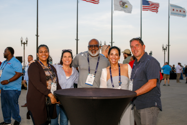 Five PWX attendees smiling around a tall table at the Get Acquainted Party. Flags fly in the background under a clear sky.