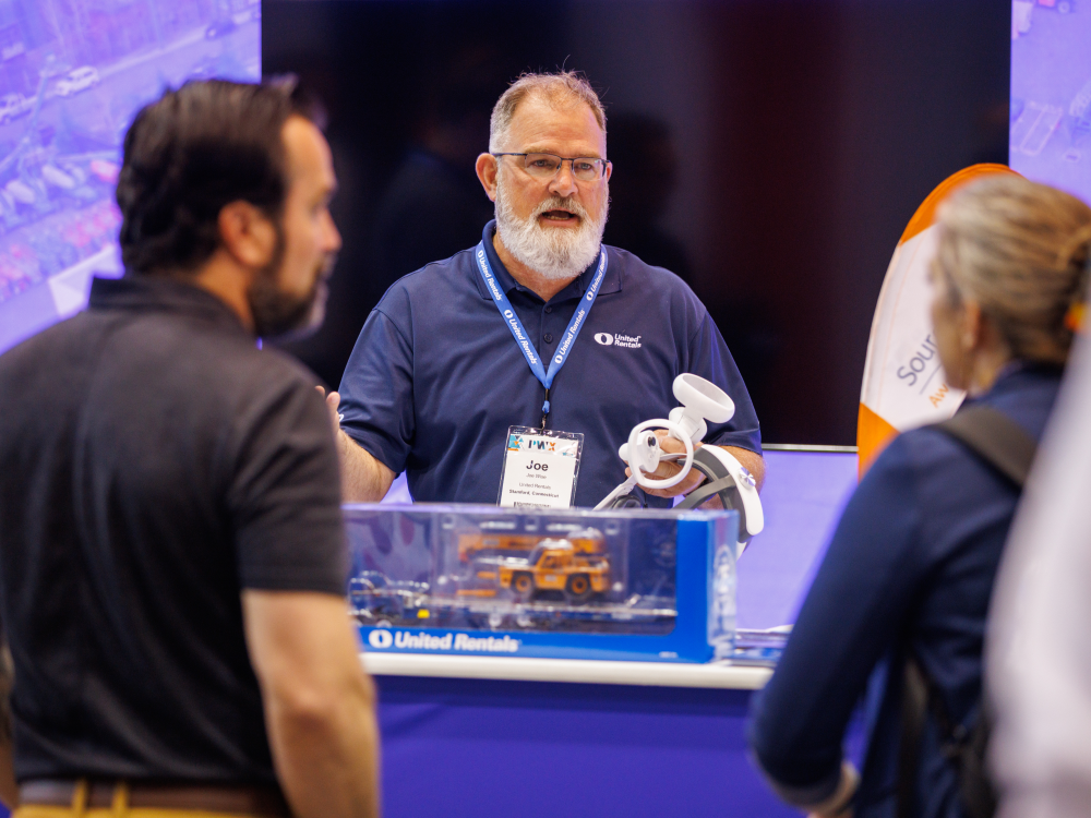A PWX exhibitor in a blue shirt talks with two attendees.