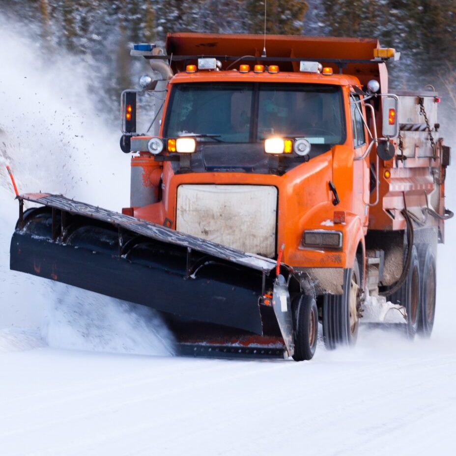 An orange truck with plow clears a road of snow.
